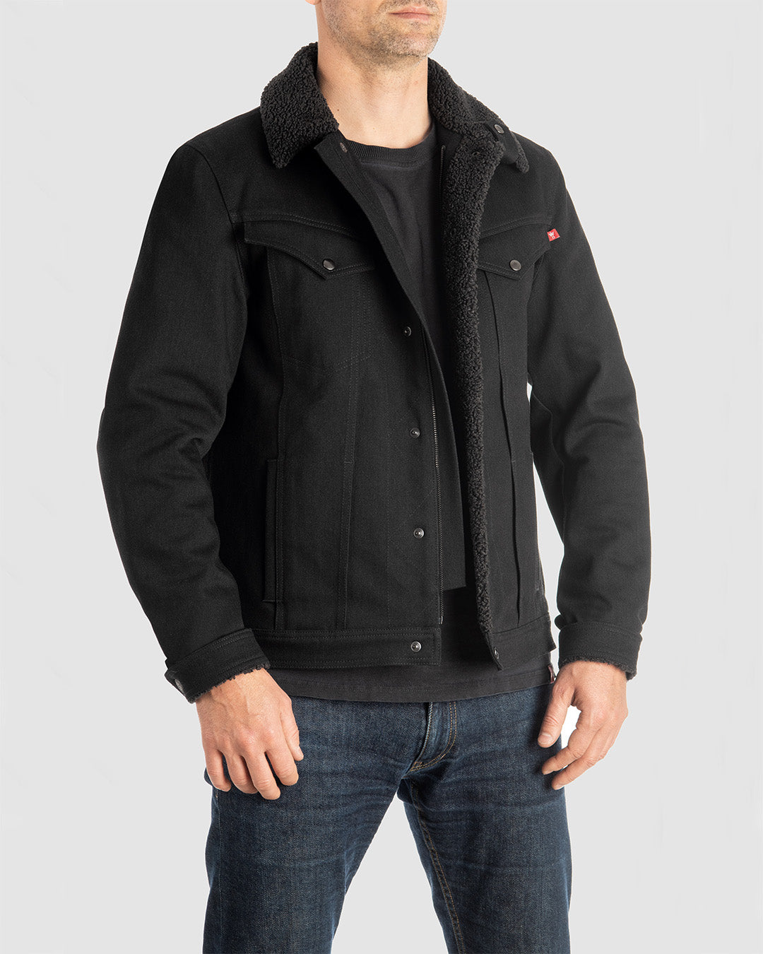 Man wearing black Husky Jacket, made with 12oz comfort-stretch CORDURA® denim, featuring Sherpa lining on the collar, cuffs, and front for warmth, and a metal YKK zipper for durable front closure.