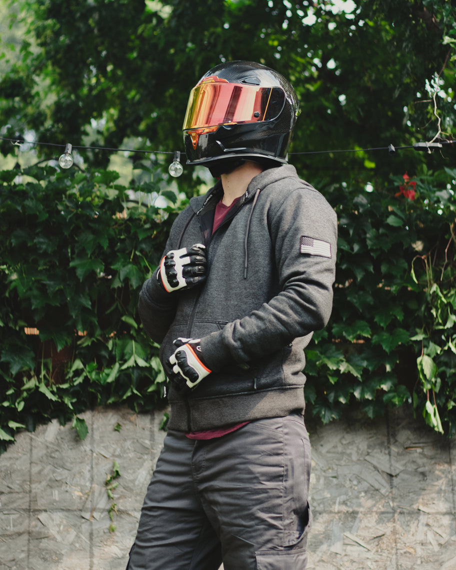Person wearing a black helmet and gray protective motorcycle hoodie standing in front of green foliage.